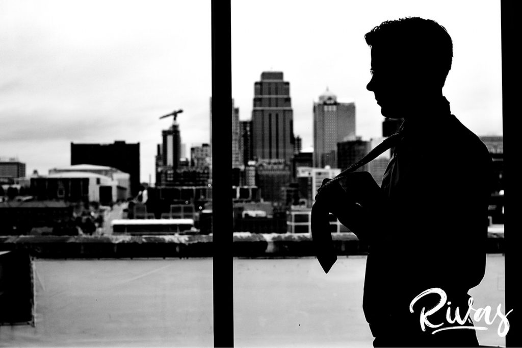 Downtown KC Library Wedding | A candid, black and white silhouette picture of a groom tying his tie on the morning of his wedding in front of the Kansas City skyline. 