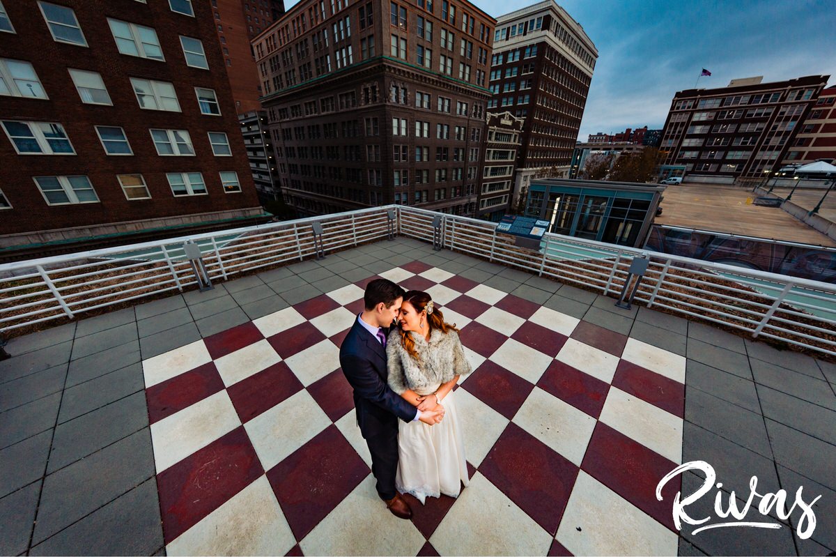 Downtown KC Library Wedding | A wide-angle, colorful portrait of a bride and groom sharing an embrace on their wedding day as they stand on the rooftop checkerboard of the downtown Kansas City library just after their first look. 