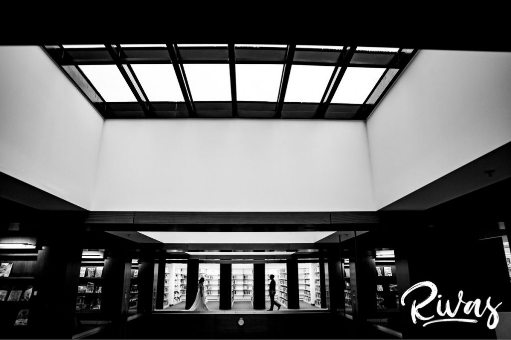 Downtown KC Library Wedding | A black and white, wide picture taken in the downtown Kansas City Public library of a bride and groom walking towards each other amongst the stacks of library books on their wedding day.