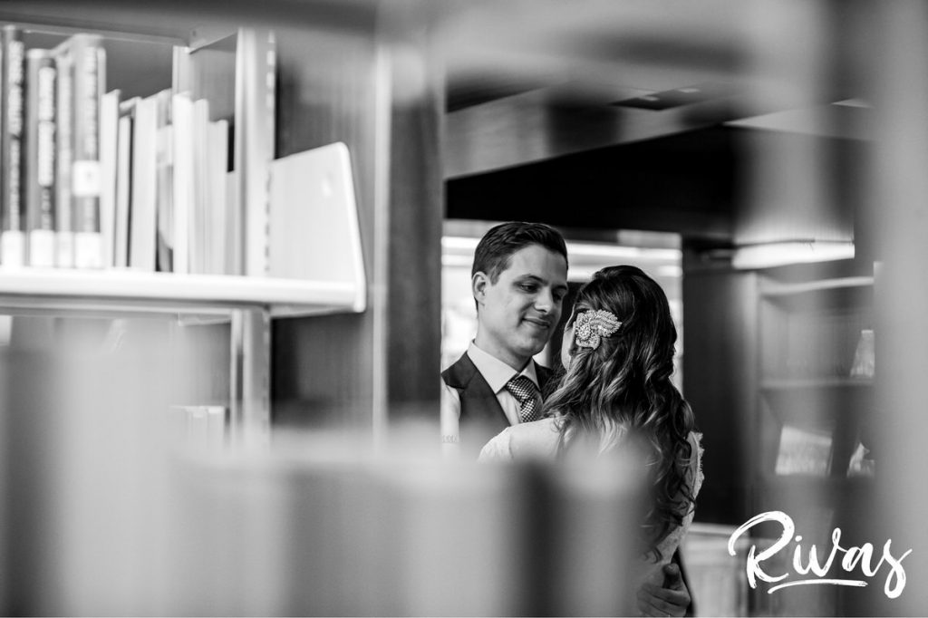 Downtown KC Library Wedding | A close-up, candid picture of a bride and groom sharing an embrace on their wedding day, taken through shelves of library books in Kansas City.
