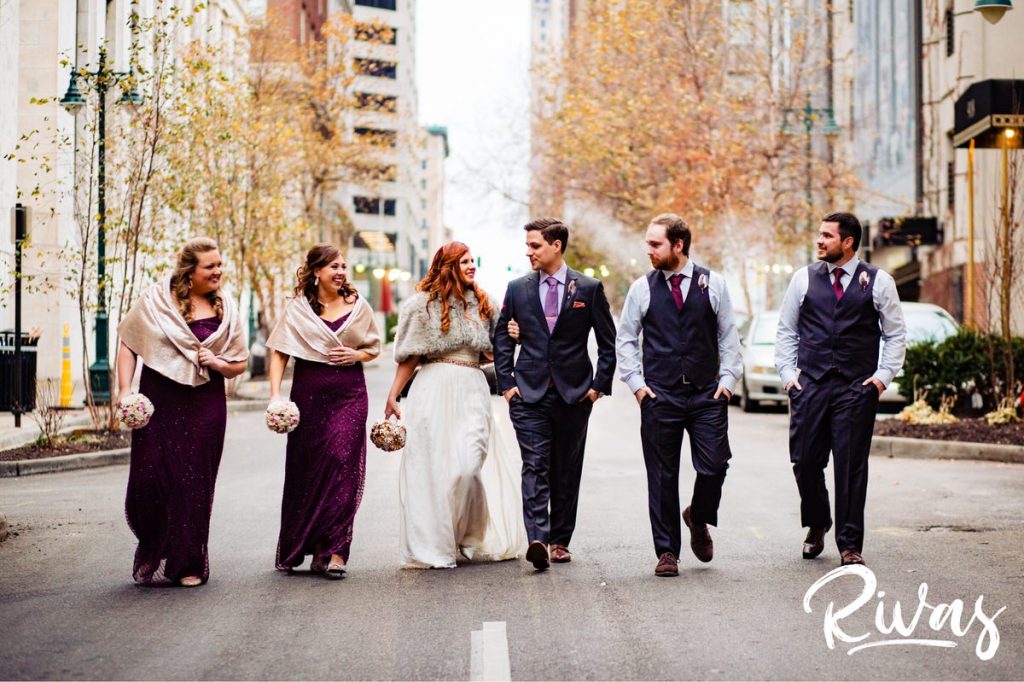 Downtown KC Library Wedding | A candid picture of a bride and groom and their wedding party dressed in plum and gray walking down Baltimore Street on their Kansas City wedding day.