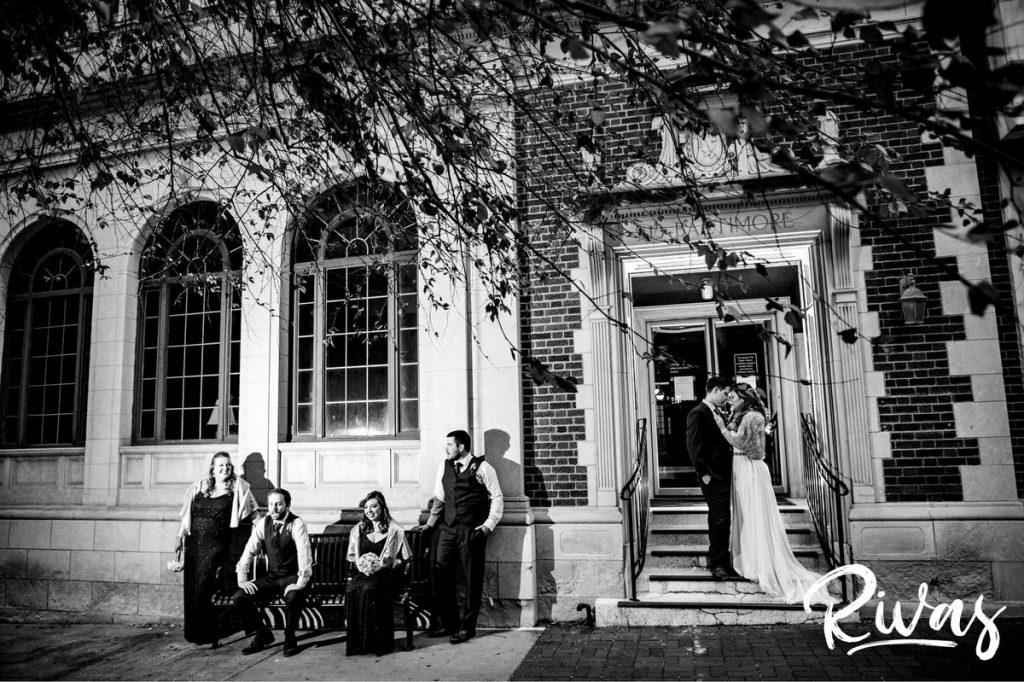 Downtown KC Library Wedding | A candid image of a bride and groom standing in a doorway as their wedding party sits on a nearby bench just outside the downtown Kansas City public library on their wedding day.