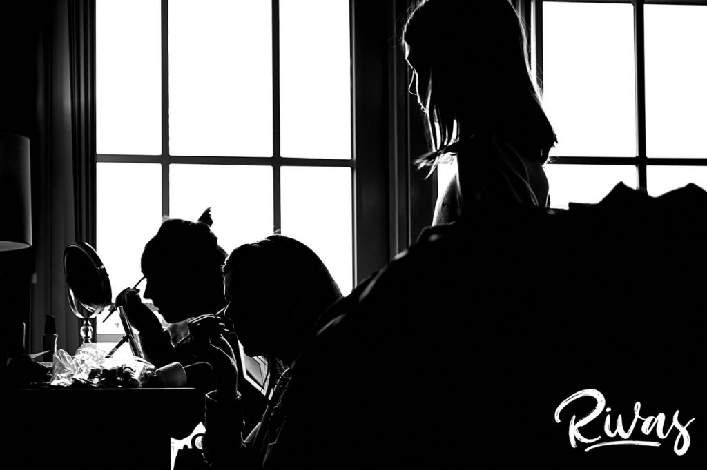 Downtown KC Library Wedding | A dramatic black and white picture of three bridesmaids sitting in front of a window putting on make-up as the flower girl watches. 