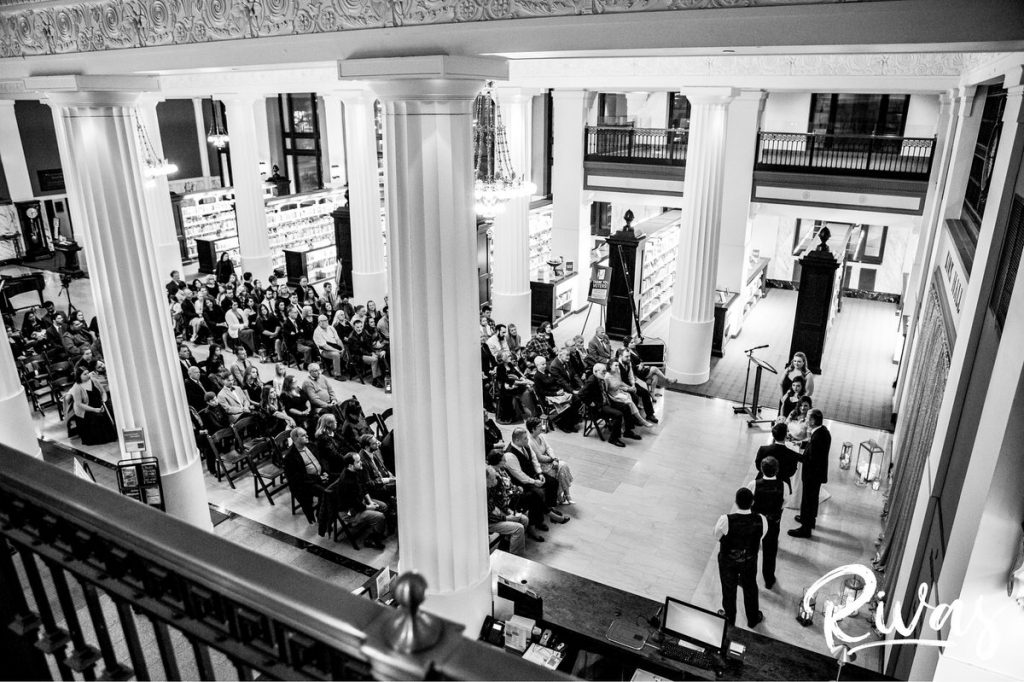 Downtown KC Library Wedding | A black and white, wide photo taken from a balcony overlooking a wedding ceremony at the Kansas City downtown public library.