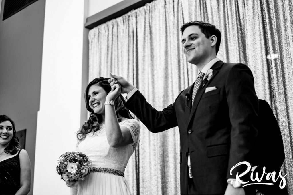 Downtown KC Library Wedding | A close-up, black and white picture of a bride and groom raising their joined hands in celebration at the end of their wedding ceremony.