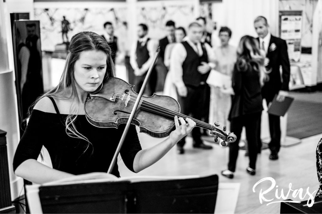 Downtown KC Library Wedding | A black and white photo of a violinist playing her violin as a bridal party lines up behind her in preparation of the wedding ceremony.