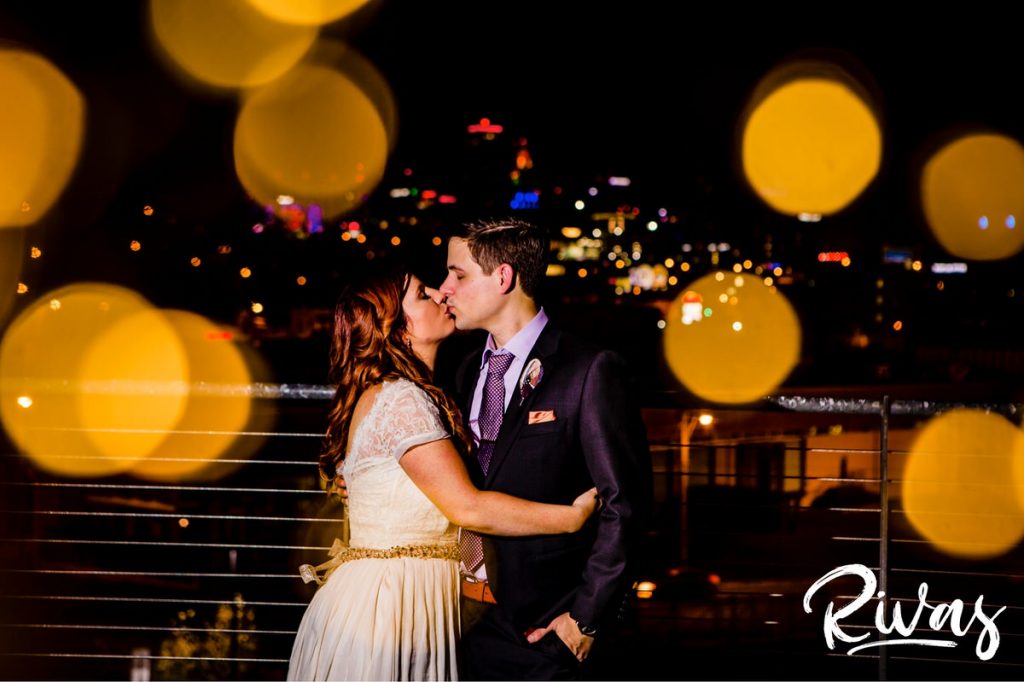 Downtown KC Library Wedding - Rivas Photography | A black and white portrait of a bride and groom as the groom kisses his bride on the forehead as they stand on the patio of the Boulevard Brewing Company in front of the Kansas City skyline during their wedding reception. 