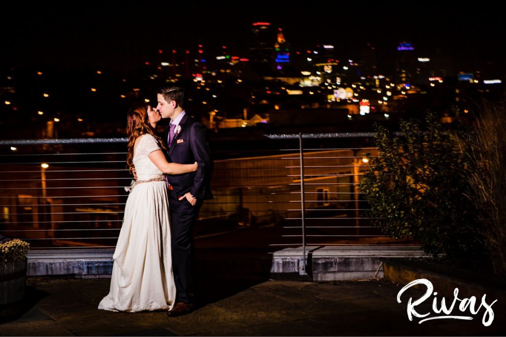 Downtown KC Library Wedding - Rivas Photography | A black and white portrait of a bride and groom as the groom kisses his bride on the forehead as they stand on the patio of the Boulevard Brewing Company in front of the Kansas City skyline during their wedding reception. 