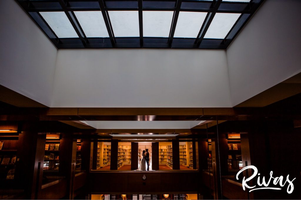 Downtown KC Library Wedding | A wide portrait of a bride and groom sharing an embrace as they stand between the stacks of library books on their wedding day at the downtown Kansas City public library.