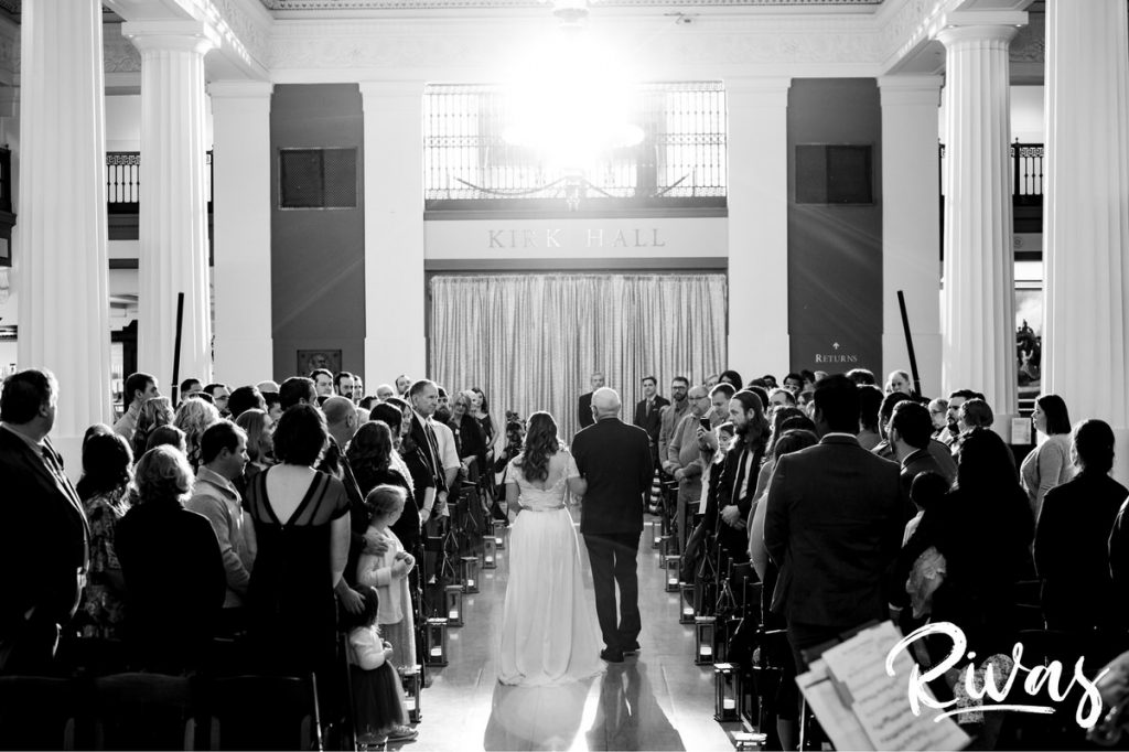 Downtown KC Library Wedding | A candid black and white picture taken from the back of the aisle of a bride and her dad walking down the aisle towards her groom as light shines down all around them.