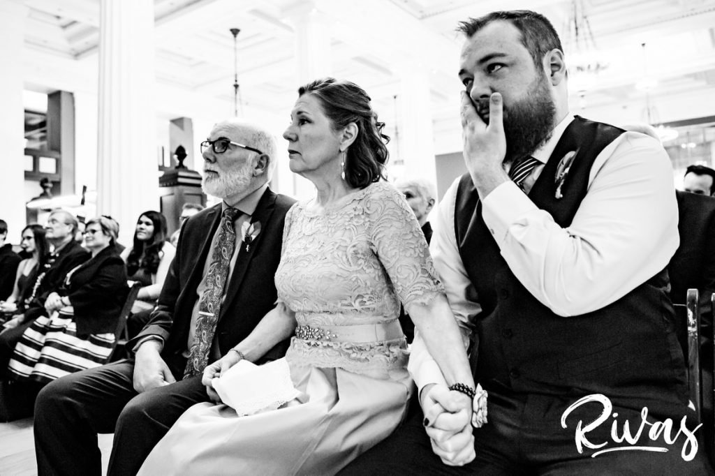 Downtown KC Library Wedding | A candid, close-up, black and white picture of a bride's mom grasping her son and husbands hand as she quietly sheds tears during her daughter's wedding ceremony.