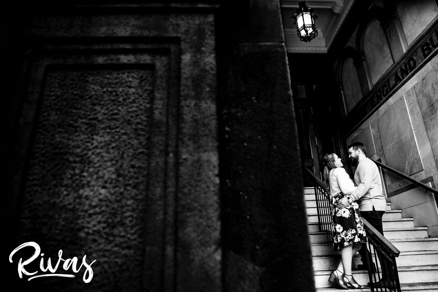 A black and white candid portrait of an engaged couple sharing an embrace as they stand in a covered staircase of a New England Building in downtown Kansas City during their engagement session. 