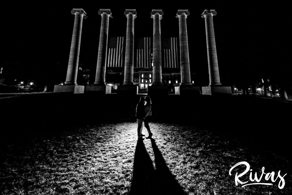 Fall MU Engagement Session A black and white, wide picture of an engaged couple sharing an embrace as they stand in front of the columns at MU after dark during their engagement session.