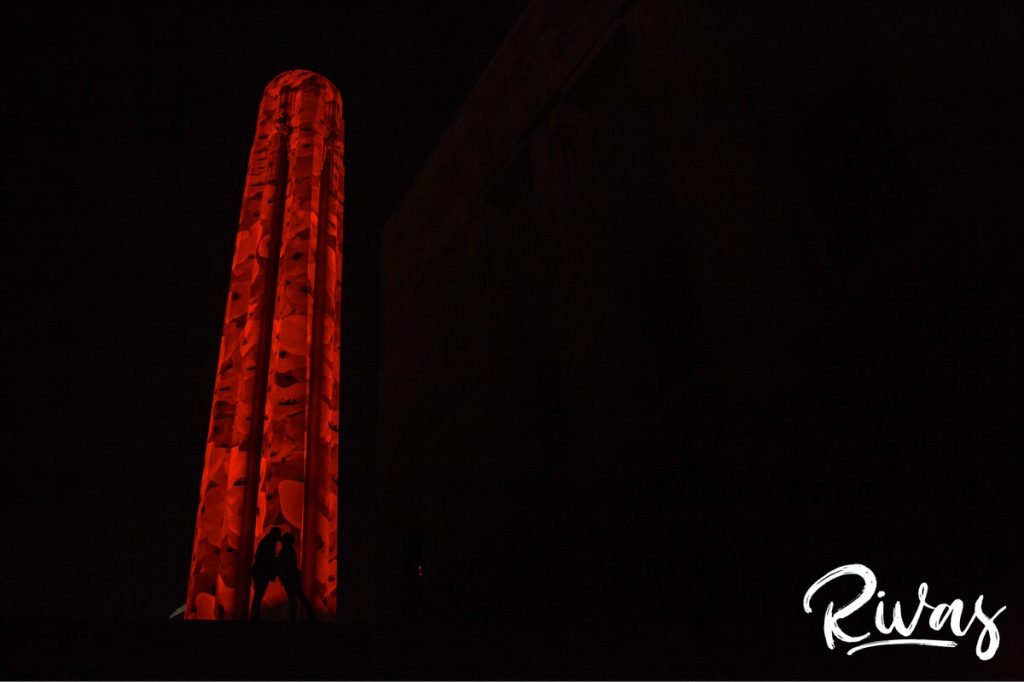 Nighttime Car Engagement Session | A silhouetted picture of an engaged couple sharing an embrace while standing in front of Kansas City's Liberty Memorial, lit up with 5000 poppies in celebration of the Centennial anniversary of the end of World War 1.
