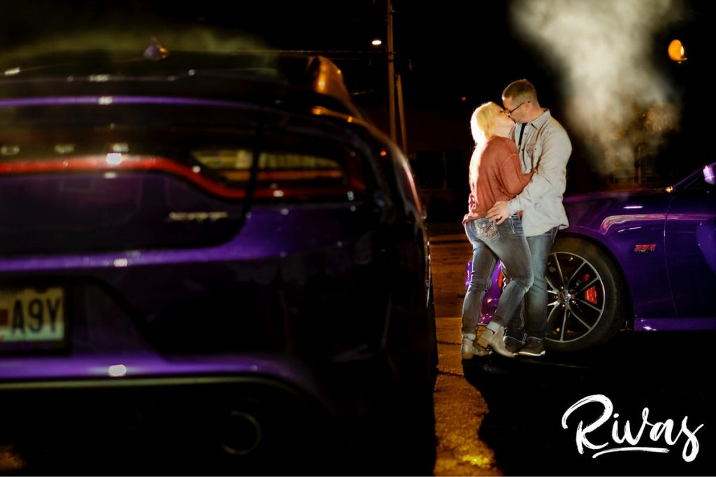 Nighttime Car Engagement Session | A dramatic nighttime portrait of an engaged couple standing back to back and holding hands in front of a pair of purple sports cars, centered in front of a Love Kansas City Mural on Southwest Boulevard.