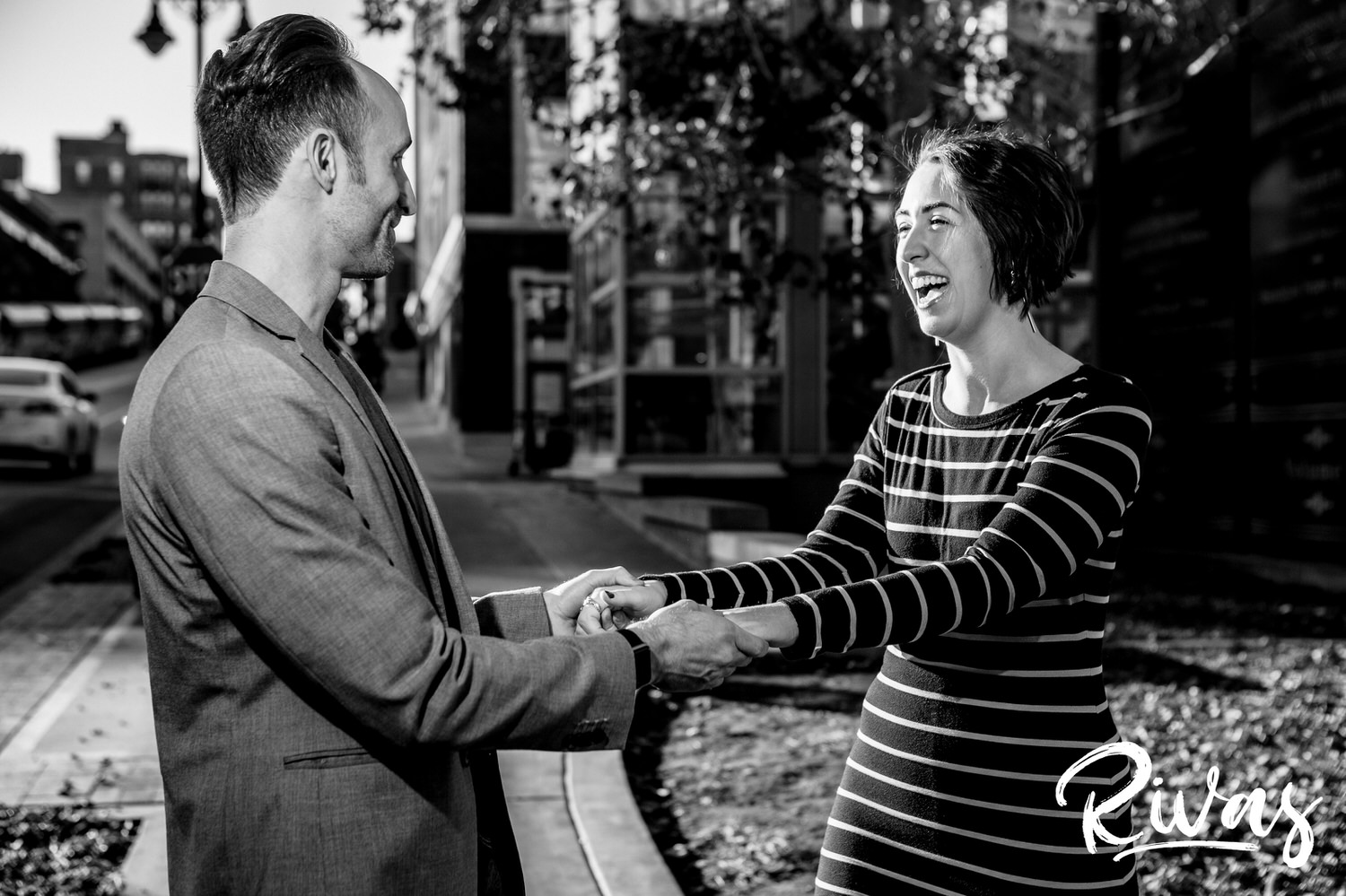 A candid black and white picture of an engaged couple holding hands, laughing and smiling together as they dance through the streets of Kansas City during their engagement session. 