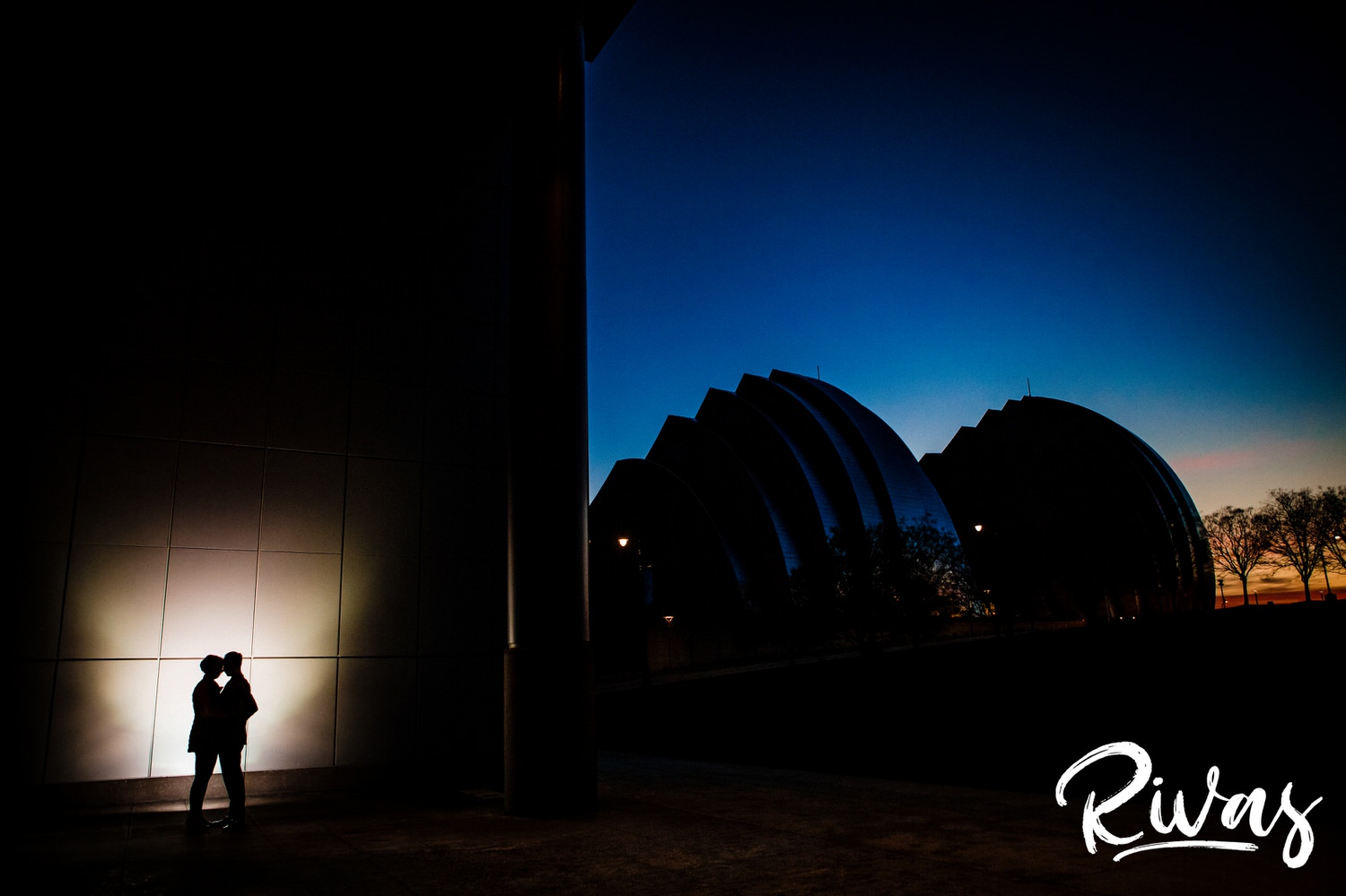 The silhouette of a couple against a steel wall with the Kauffman Center for the Performing Arts visible in the background at sunset during their engagement session. 