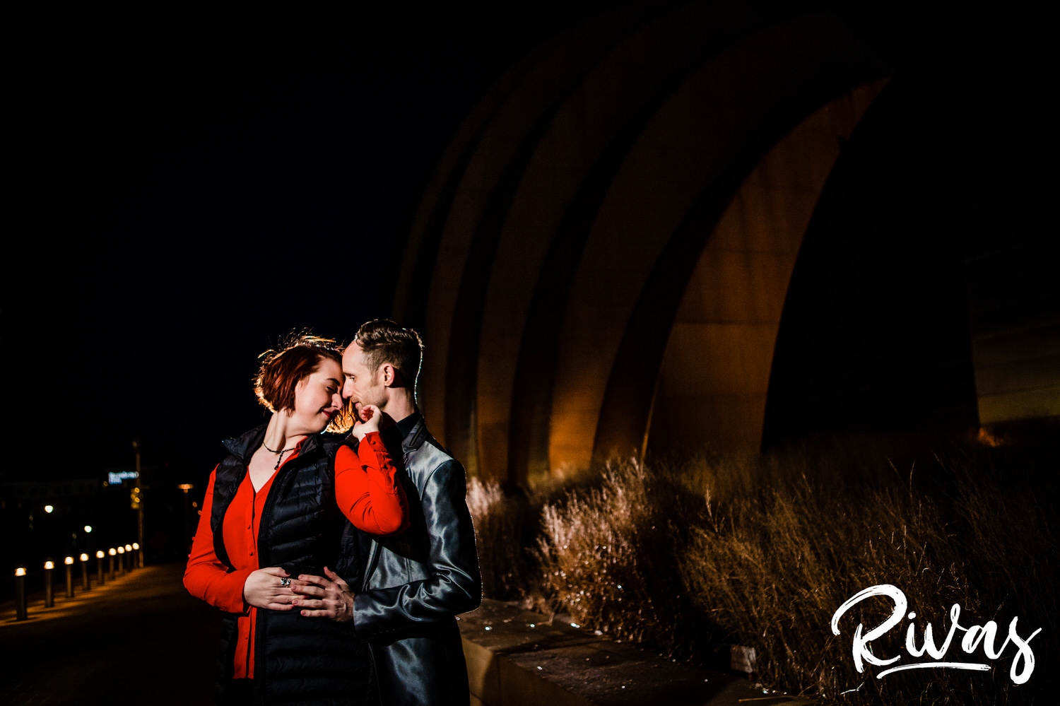 An intimate picture of an engaged couple sharing an embrace outside the Kauffman Center for the Performing Arts in Kansas City after dark. 