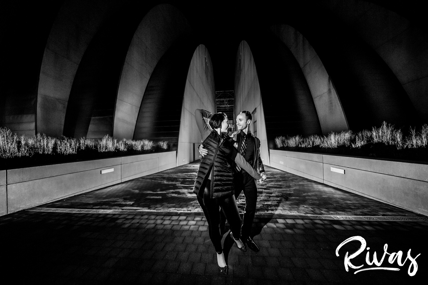 A black and white picture of an engaged couple dancing in the dark in front of the Kauffman Center for the Performing Arts during their engagement session in Kansas City. 
