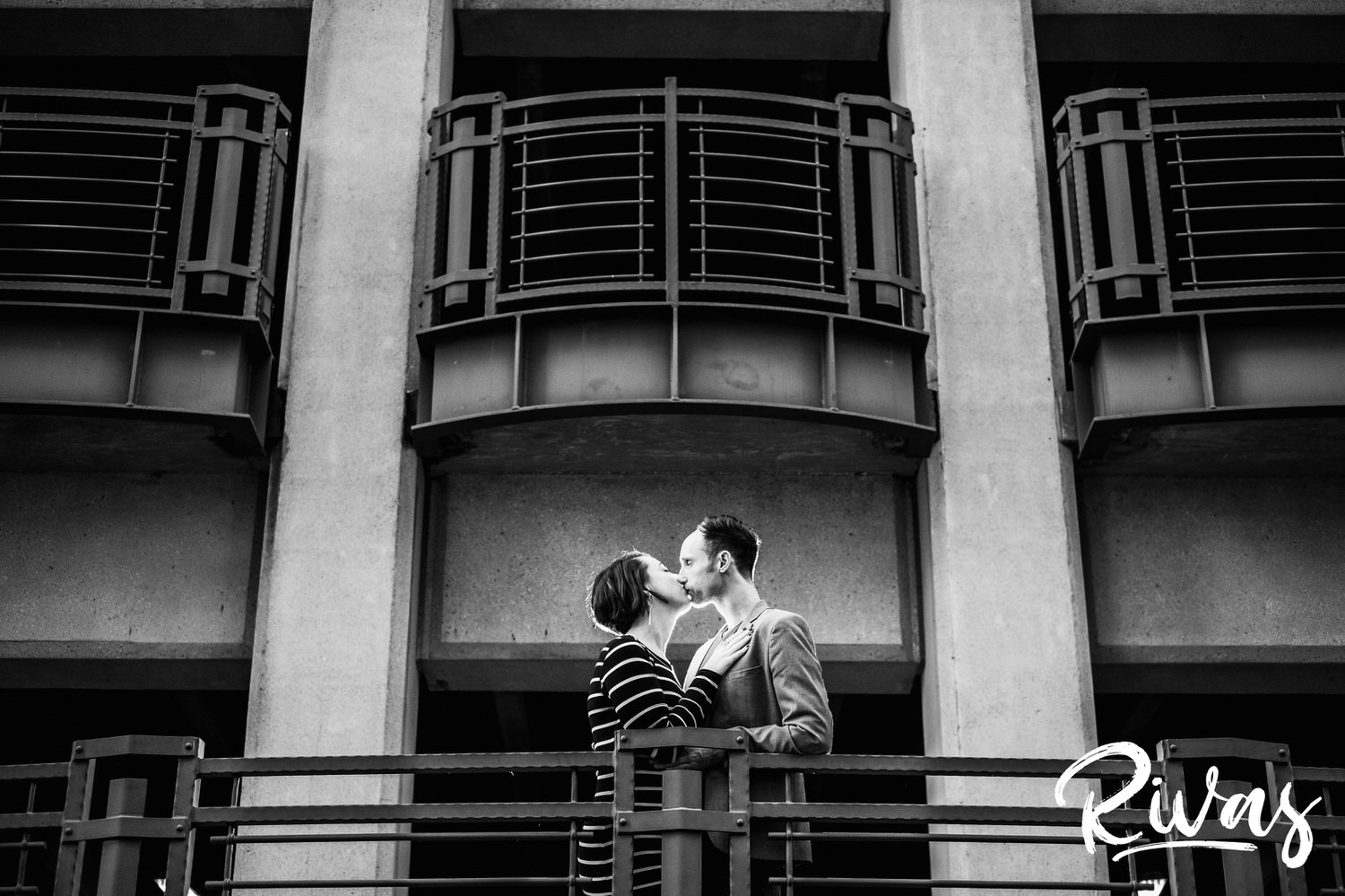 A black and white portrait of an engaged couple standing on the portico of the downtown Kansas City Library parking garage, embracing and sharing a kiss. 