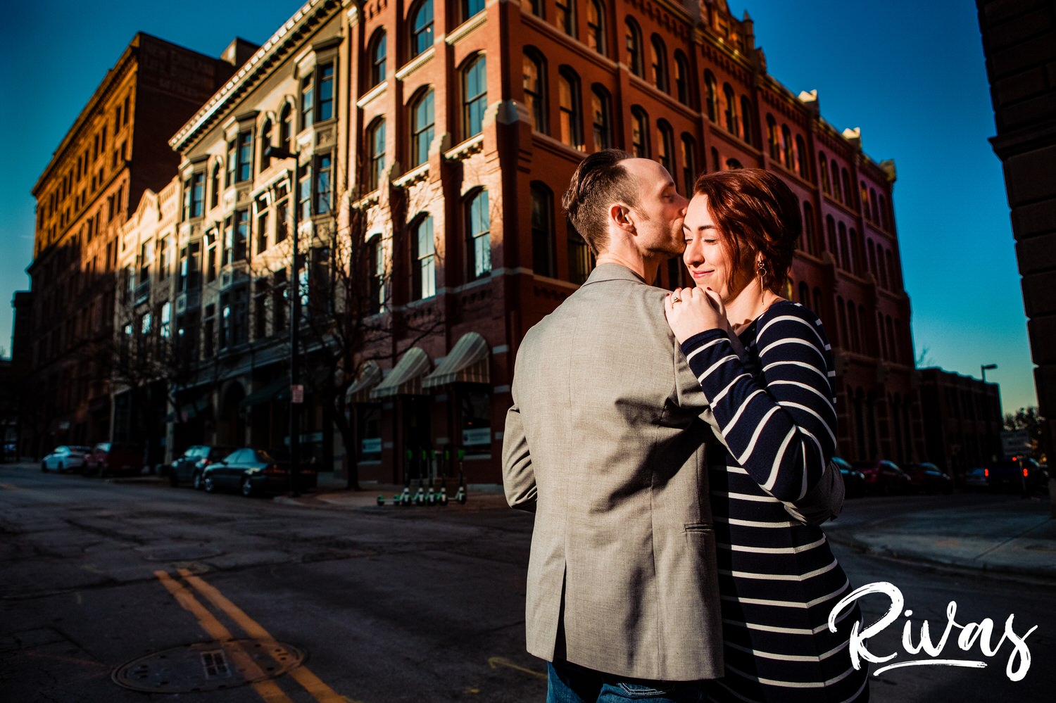 A vibrant, candid picture of an engaged couple walking down the street as he kisses her on the forehead during their engagement session in Kansas City. 