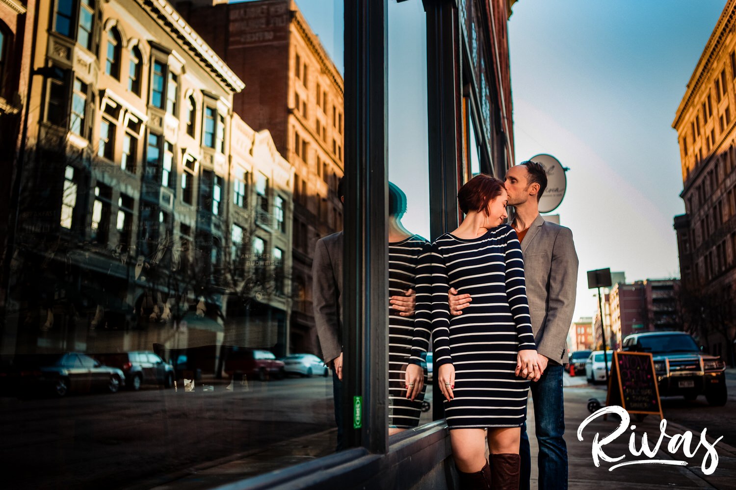 A portrait of an engaged couple embracing as they stand against the glass of a deli in downtown Kansas City during their engagement session. 