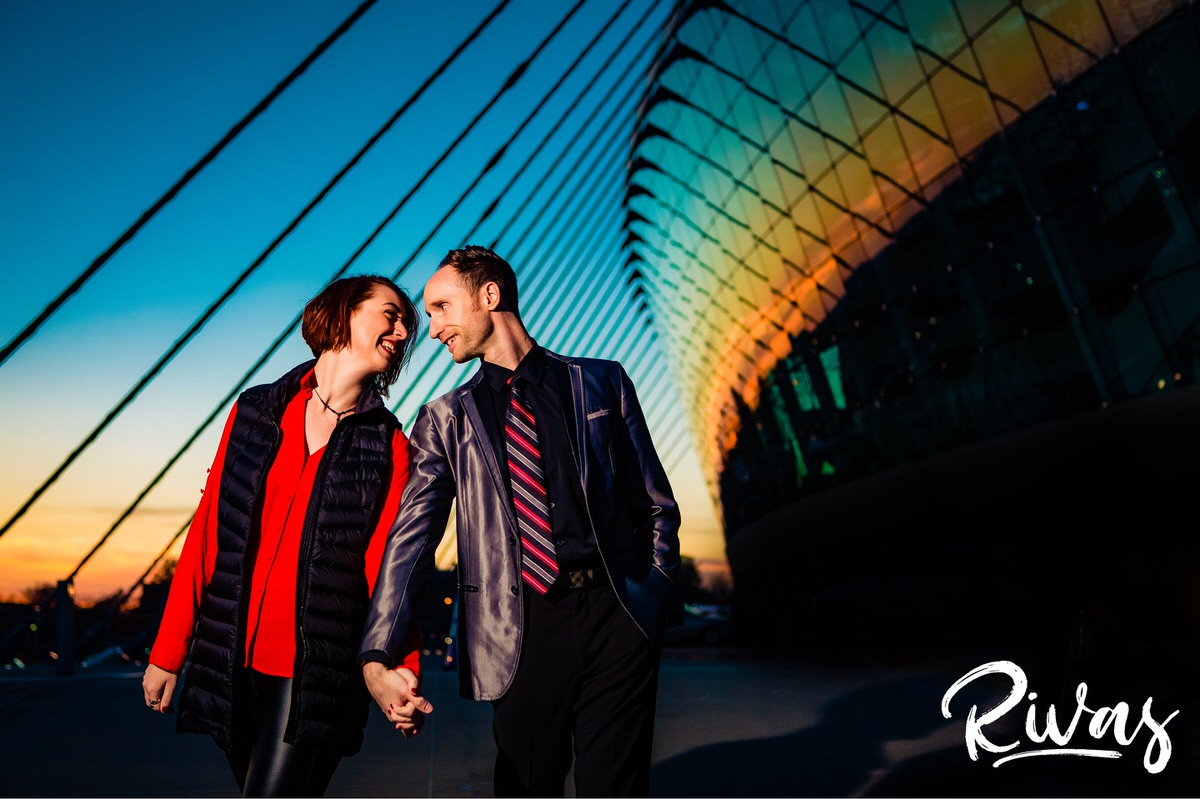 A candid picture of an engaged couple holding hands and walking towards the camera in front of the Kauffman Center for the Performing arts in Kansas City at sunset. 