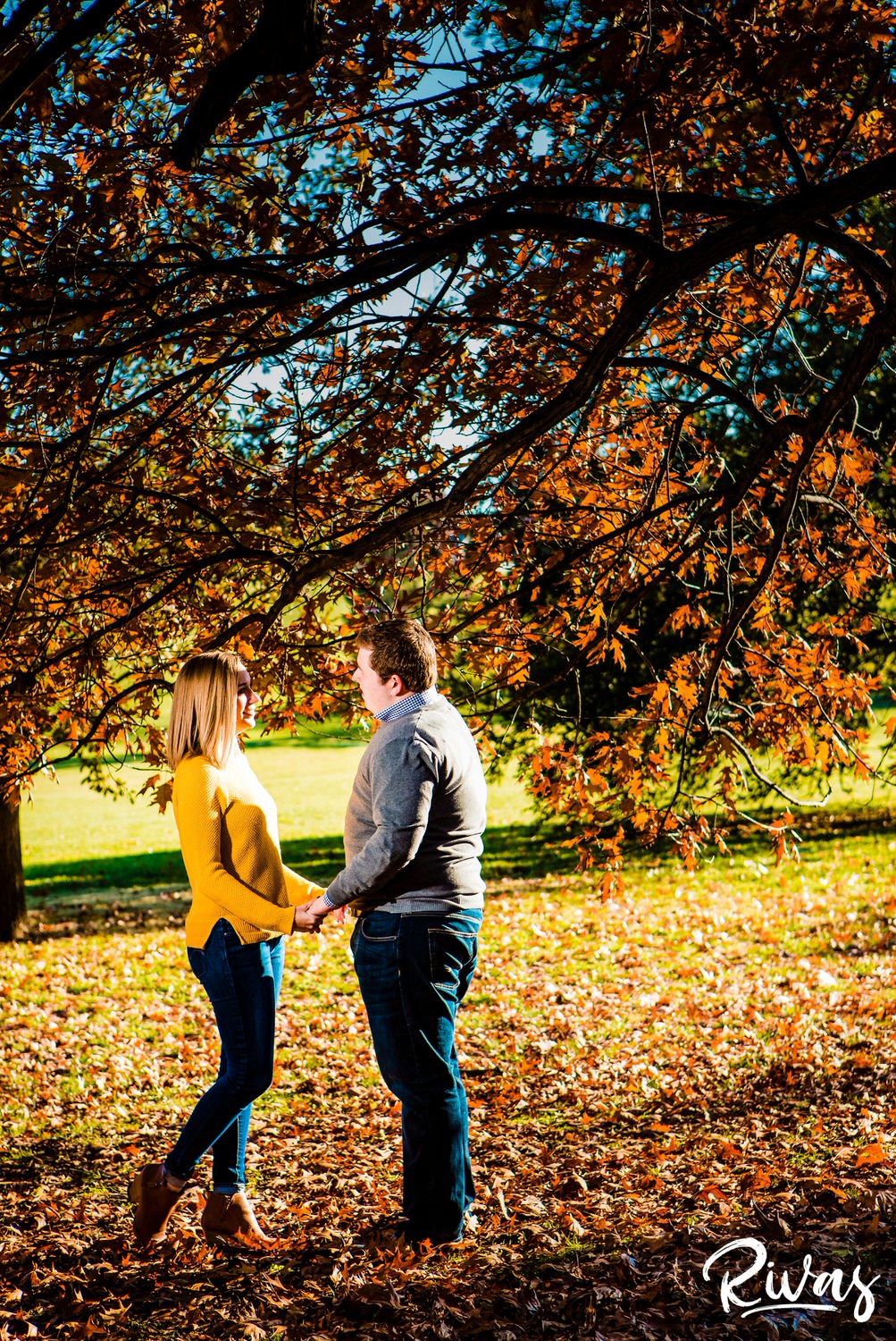 A candid picture of an engaged couple holding hands and smiling at each as they stand on a bed of bright orange leaves and under a tree covered in orange leaves during their engagement session at Kansas City's Loose Park. 