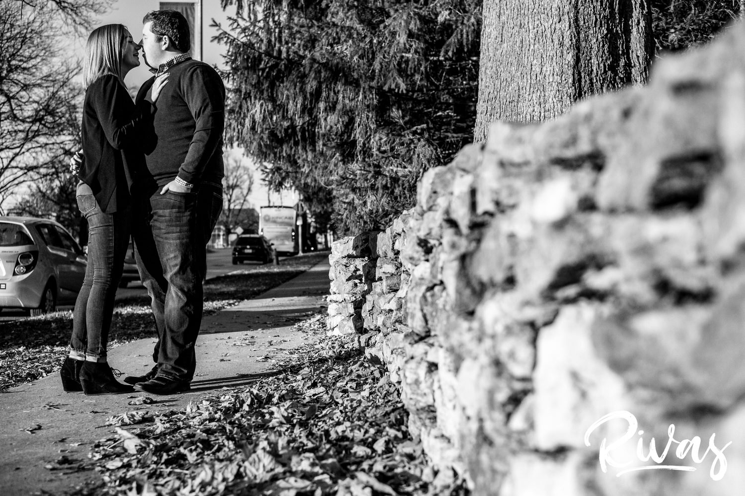 A black and white portrait of a couple stopping to share a kiss on a sidewalk outside The Nelson during their engagement session. 
