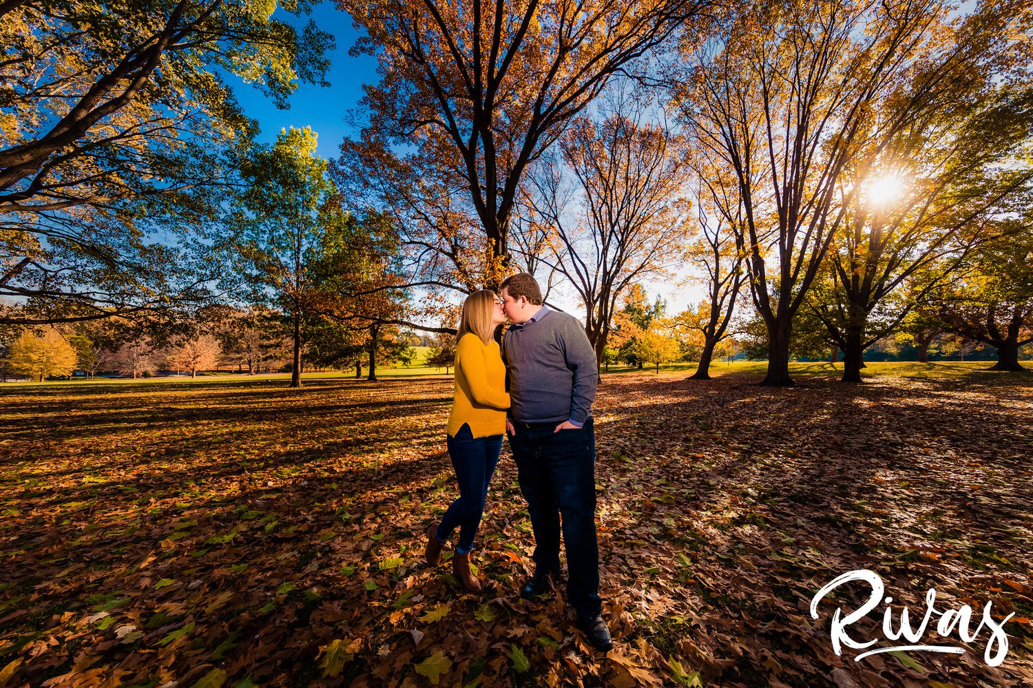 A vibrant picture of an engaged couple embracing and sharing a kiss as they stand under a colorful canopy of fall leaves at Loose Park during their engagement session in Kansas City. 