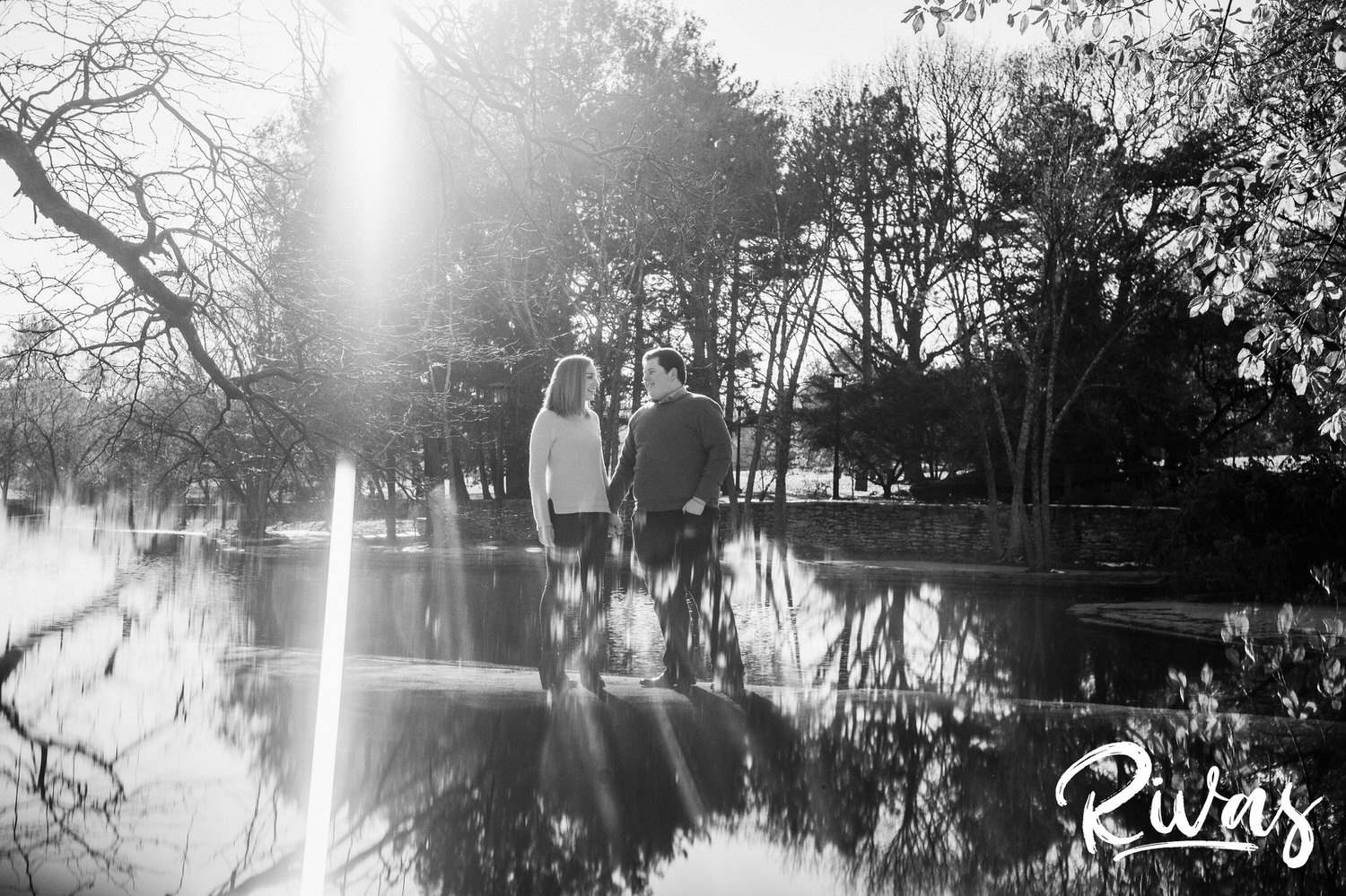 A black and white picture of an engaged couple holding hands as they stand on a cement bridge going over the center of the pond at Loose Park in Kansas City. 