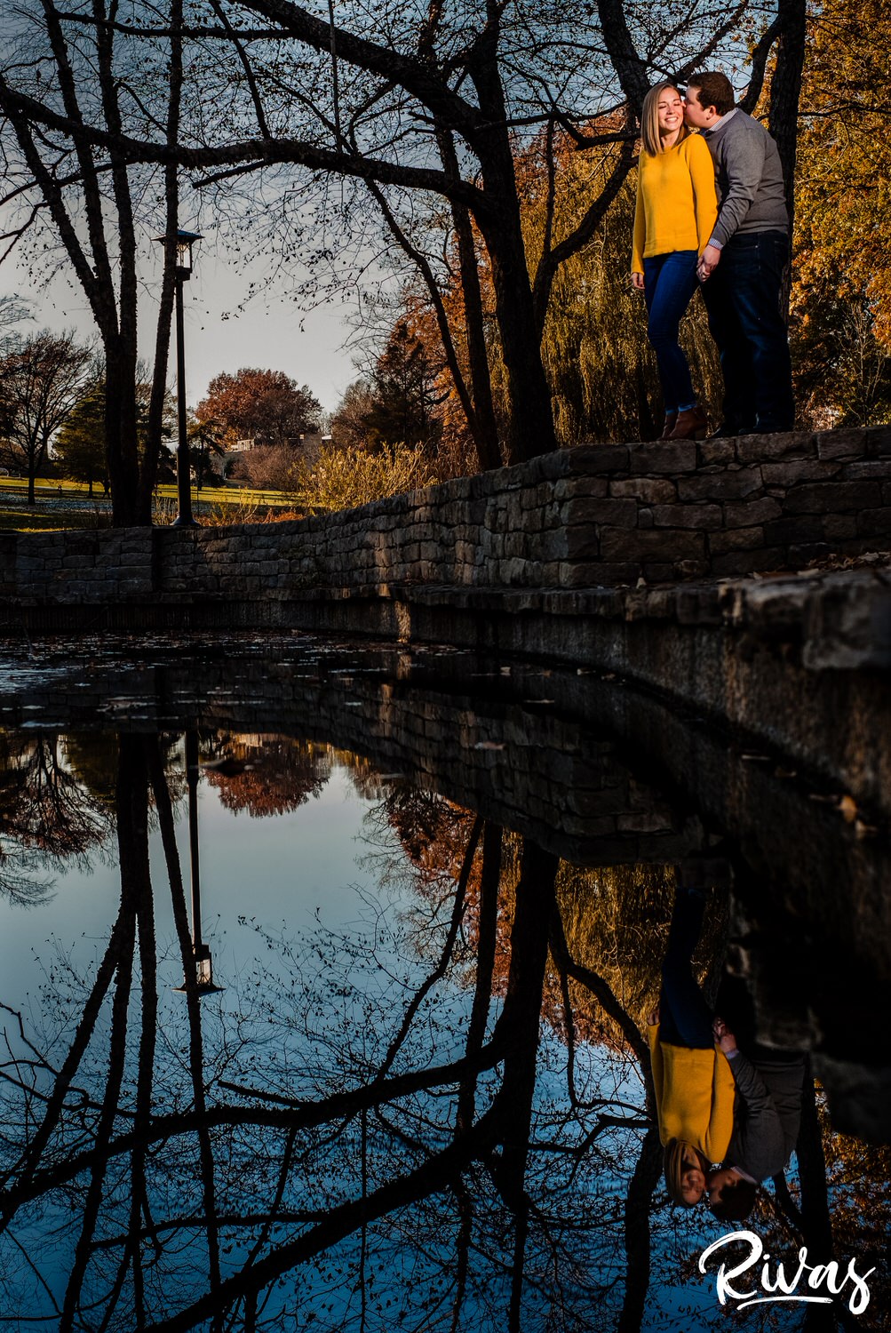 A portrait of an engaged couple, standing together on the edge of a pond sharing an embrace while the water below them reflects their image during their engagement session at Loose Park. 