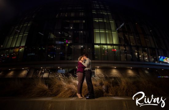 Winter Kauffman E-Session Sneak Peek | A portrait of an engaged couple embracing and sharing a kiss while standing outside Kansas City's Kauffman Center during their night-time engagement session.