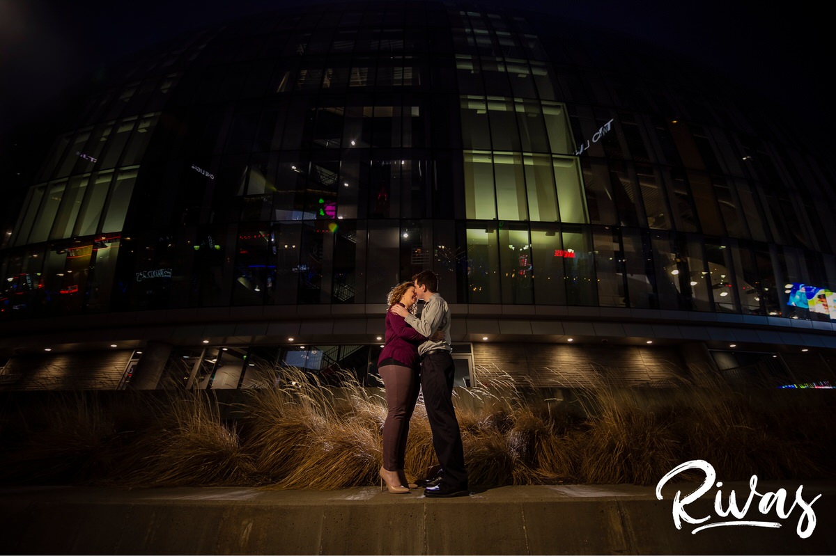 Winter Kauffman E-Session Sneak Peek | A portrait of an engaged couple embracing and sharing a kiss while standing outside Kansas City's Kauffman Center during their night-time engagement session.