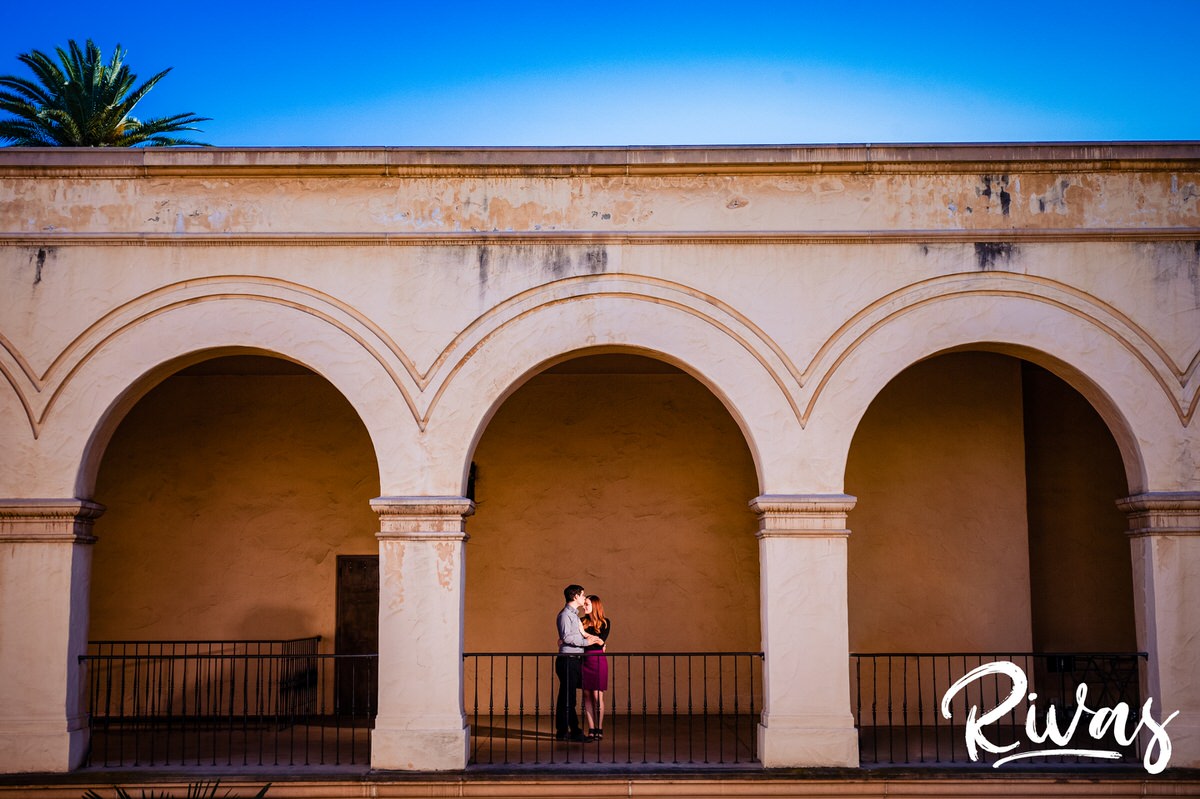 San Diego Destination Engagement Session | A portrait of an engaged couple sharing an embrace while standing underneath a series of arches with a peek of blue sky and palm tree above them.