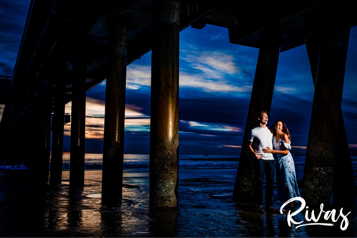 Vibrant Venice Engagement Session | A candid image of an engaged couple embracing and laughing while standing underneath the pier at Venice Beach.
