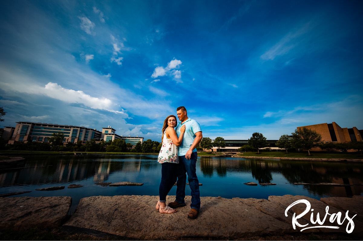 Summer Morning Session Sneak Peek | A vibrant photo of an engaged couple standing on a rock wall in front of a vibrant blue pond and sky near Kansas City's Plaza Neighborhood.