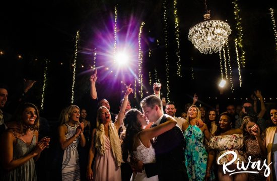 Destination Wedding Photographer | Saddlerock Ranch Summer Wedding | A candid picture of a bride and groom embracing underneath a canopy of twinkling lights and chandeliers surrounded by family and friends on the dancefloor during their wedding reception at Saddlerock Ranch in Malibu, California.