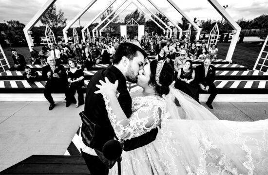 A candid, black and white picture taken from behind the bride and groom, looking out at their audience as they share their first kiss on their wedding day at The Pavilion in Kansas City.