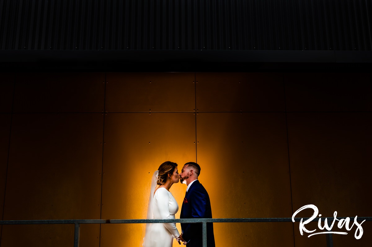 Kansas City Urban Wedding Sneak Peek | A back, spotlit image of a bride and groom standing in front of a deep gold wall sharing a kiss on their wedding day in downtown Kansas City.