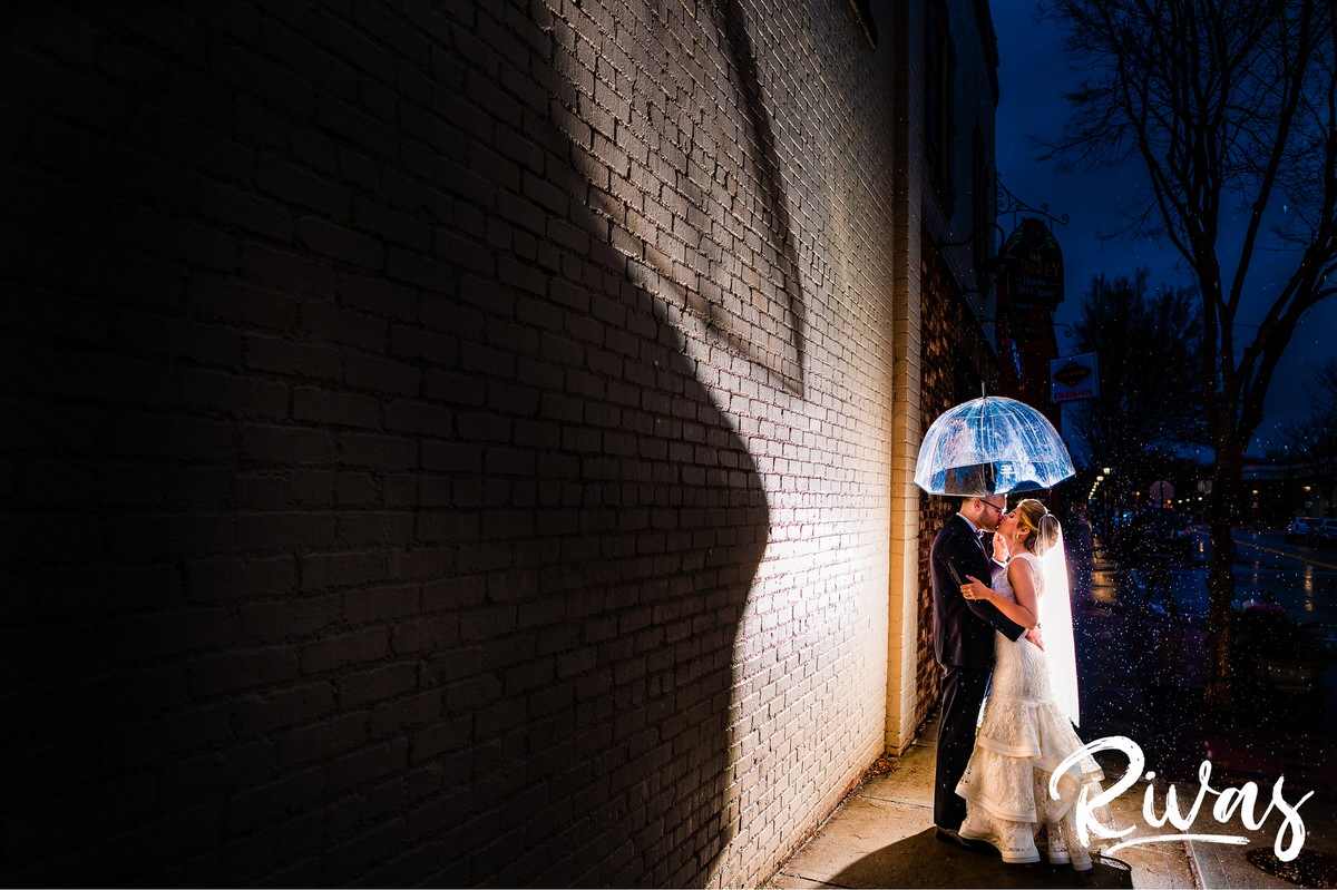 Downtown Lee's Summit Wedding Sneak Peek | A dramatic night time picture of a bride and groom standing next to a cream brick wall underneath a clear umbrella as the rain pours down around them on their wedding day at The Stanley Event Space in downtown Lee's Summit.
