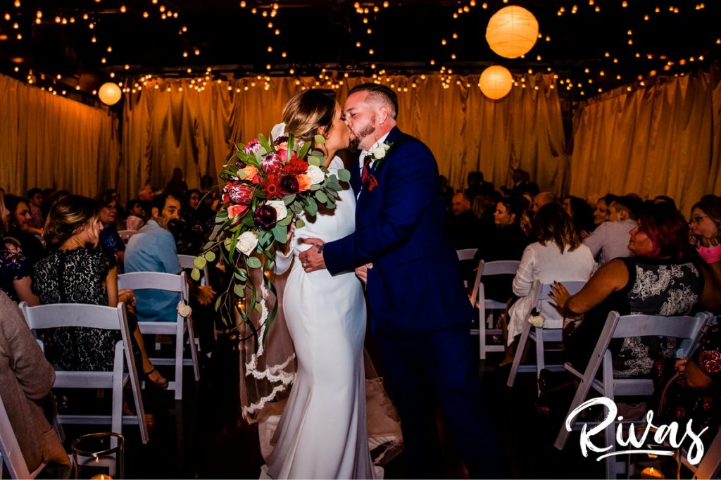 A colorful image of a bride and groom sharing a kiss at the end of the aisle just after their wedding ceremony at 28 Event Space in Kansas City. | Kansas City Room Wedding & Reception | Rivas Weddings