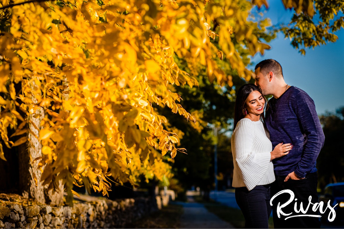 Colorful Nelson Engagement Session | A candid picture of an engaged couple sharing an embrace as they stand in front of a tree with vibrant yellow leaves during their engagement session at The Nelson Atkins Museum of Art in Kansas City.