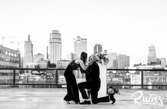 KC Skyline Proposal | A black and white, wide picture of a man down on one knee proposing to his girlfriend as she leans in to kiss him after saying yes.