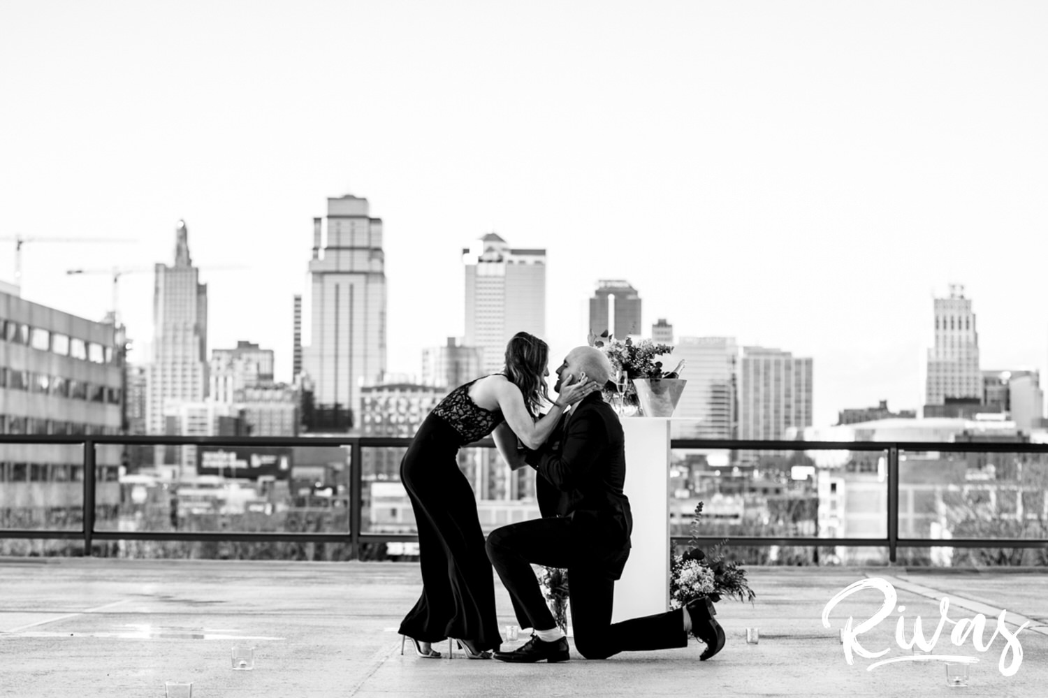 KC Skyline Proposal | A black and white, wide picture of a man down on one knee proposing to his girlfriend as she leans in to kiss him after saying yes.