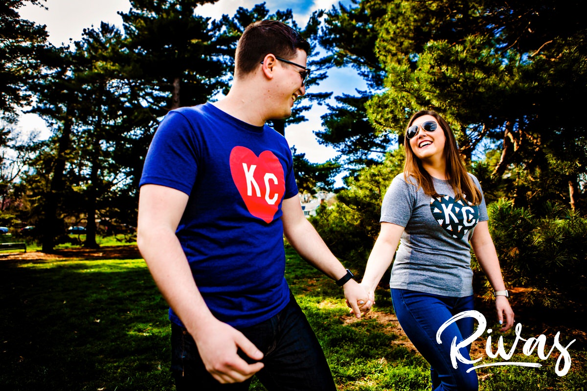 A colorful, candid picture of a couple in jeans and Charlie Hustle tee-shirts holding hands and walking through Loose Park during their spring engagement session in Kansas City. 