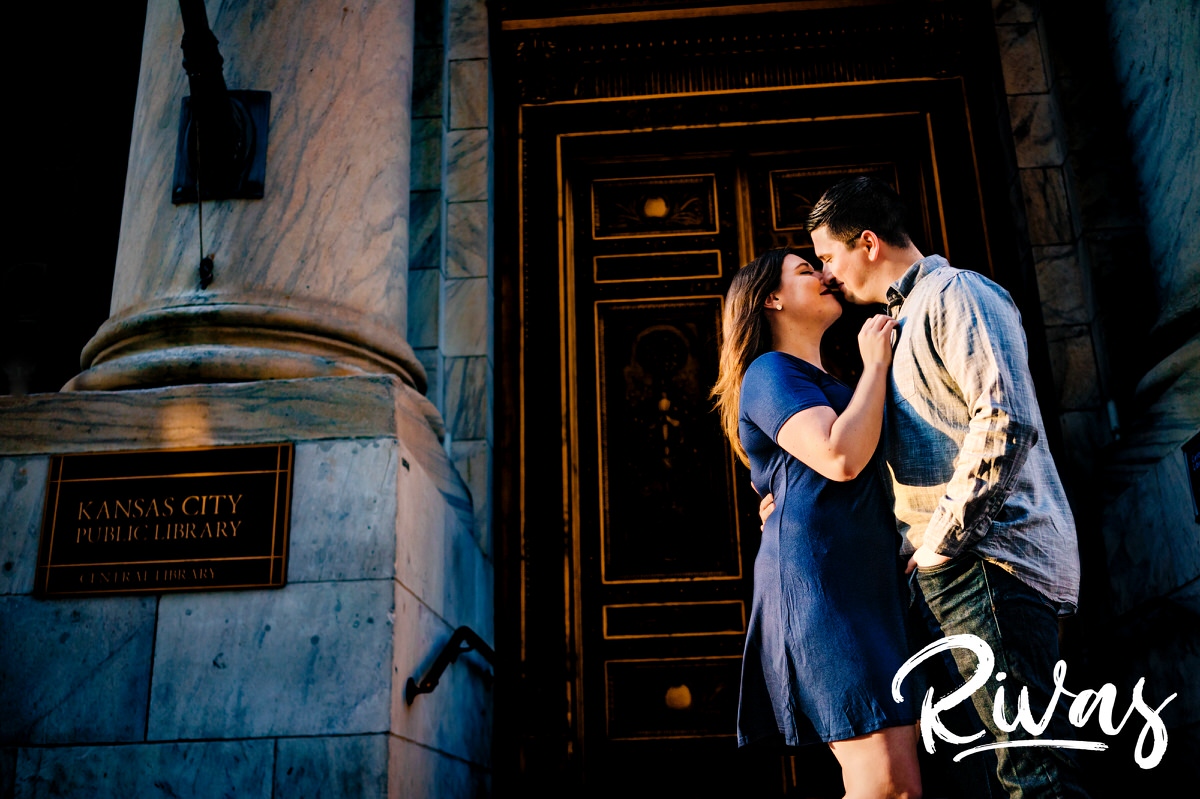 A candid picture of an engaged couple sharing a kiss as they stand in front of the downtown Kansas City Library at sunset during their spring engagement session. 