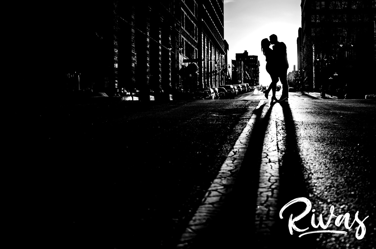 A dramatic black and white picture of an engaged couple standing in the middle of the street and sharing a kiss outside the central branch of the downtown public library in Kansas City during their engagement session. 
