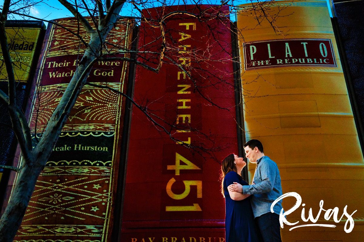 A colorful picture of an engaged couple sharing an embrace as they stand in front of the downtown Kansas City Library parking garage, covered with book spines during their spring engagement session. 