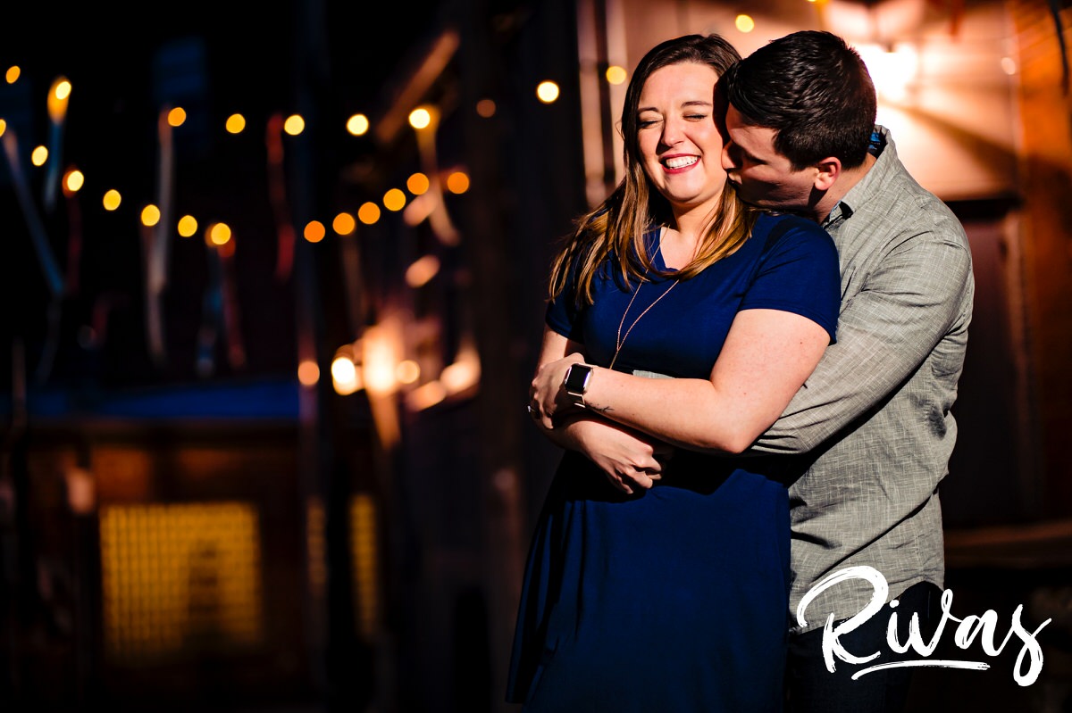 A candid, vibrant picture of an engaged couple sharing an embrace and laughing together under twinkly lights hung in an alley in Kansas City's Crossroads District. 